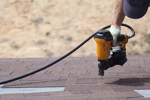 Roofer builder worker with nailgun installing Asphalt Shingles or Bitumen Tiles on a new house under construction